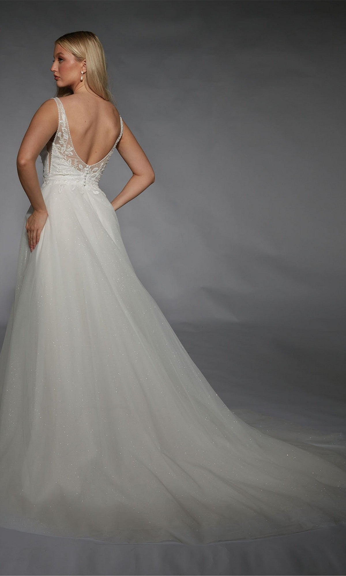 A woman stands facing away in front of a gray background, wearing the Long White Dress: Alyce 7125—a sleeveless gown with a sheer lace bodice and a flowing train, radiating timeless elegance.